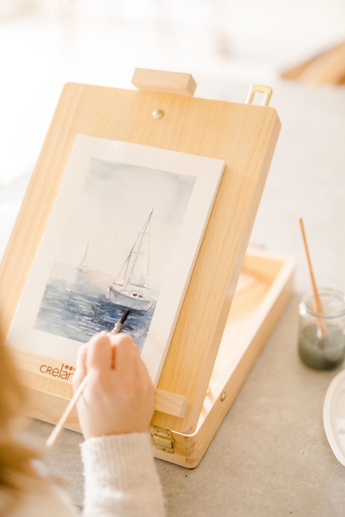 Atelier artisanat Vendée : main peignant une aquarelle de bateau sur chevalet dans une ambiance douce et créative