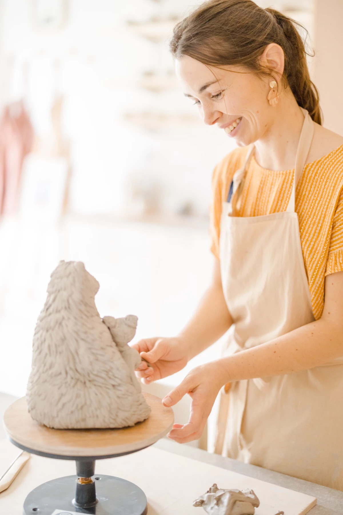 Portrait corporate artisan : céramiste souriante travaillant une sculpture d'ours en terre dans son atelier lumineux en Vendée