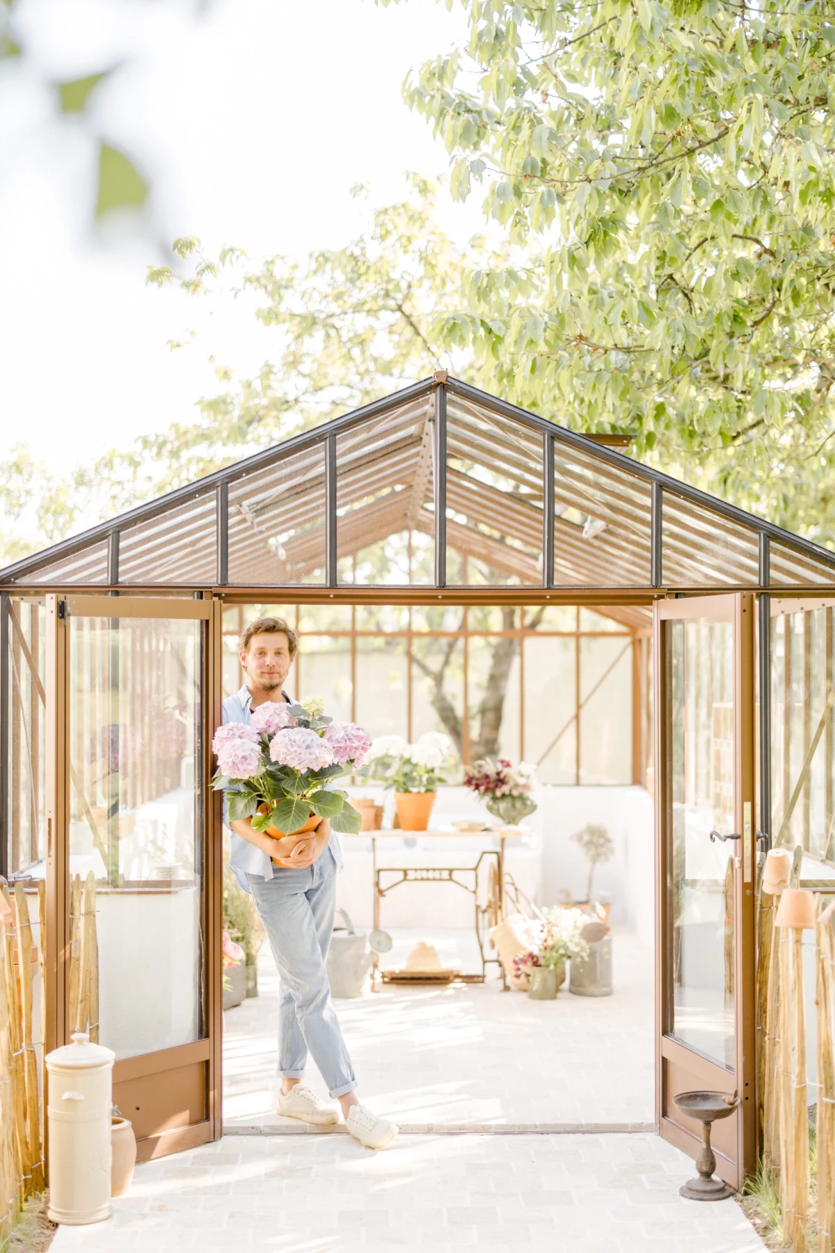 Portrait corporate fleuriste : artisane en tenue claire porte un bouquet de fleurs pastel dans une verrière baignée de lumière naturelle