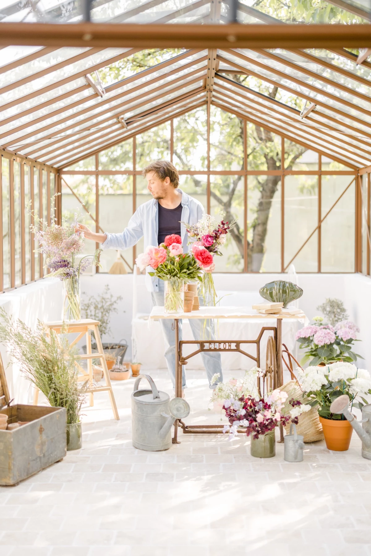 Portrait corporate fleuriste dans verrière lumineuse, arrangeant bouquets champêtres aux tons roses et blancs, ambiance douce et professionnelle