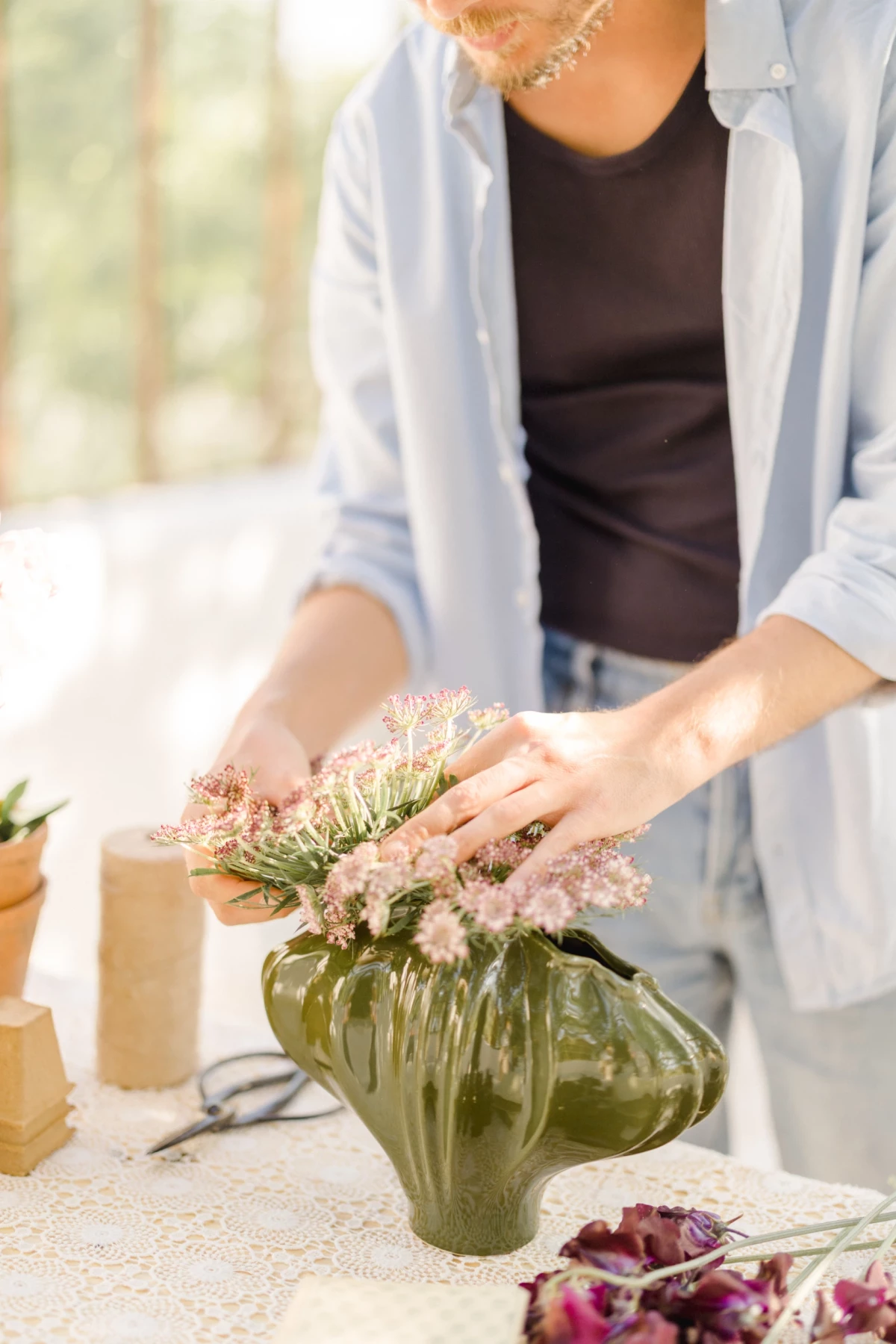 Séance corporate fleuriste : mains arrangeant délicatement des fleurs séchées roses dans un vase vert, ambiance douce et artisanale
