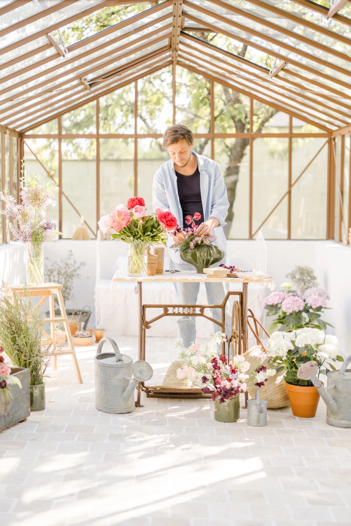 Portrait corporate fleuriste : femme arrange un bouquet de pivoines roses dans une verrière baignée de lumière naturelle douce