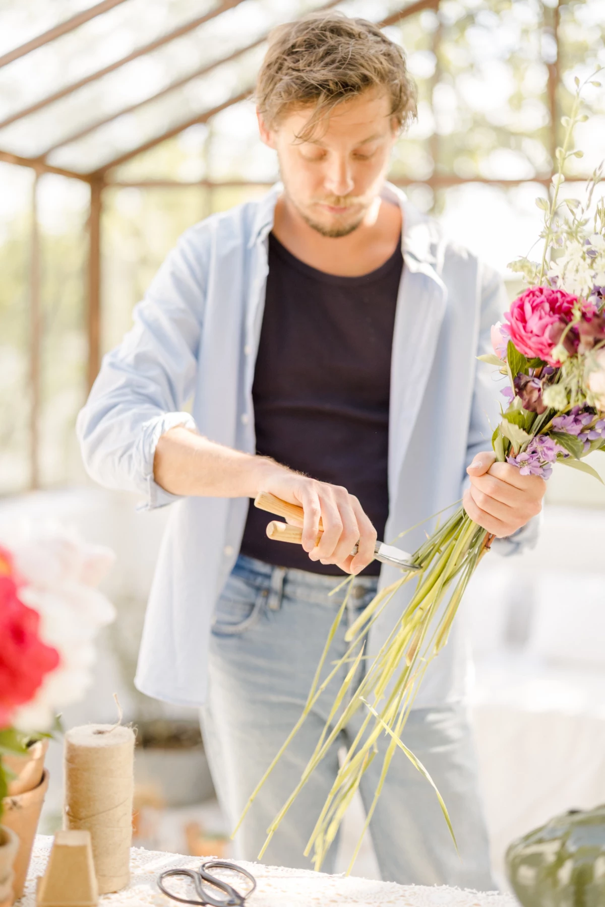 Portrait corporate fleuriste : professionnel créant un bouquet de fleurs roses dans une verrière lumineuse, geste délicat et savoir-faire