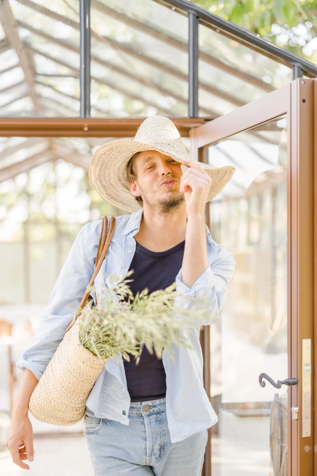 Portrait corporate fleuriste souriant sous chapeau de paille avec bouquet, ambiance douce et lumineuse devant verrière