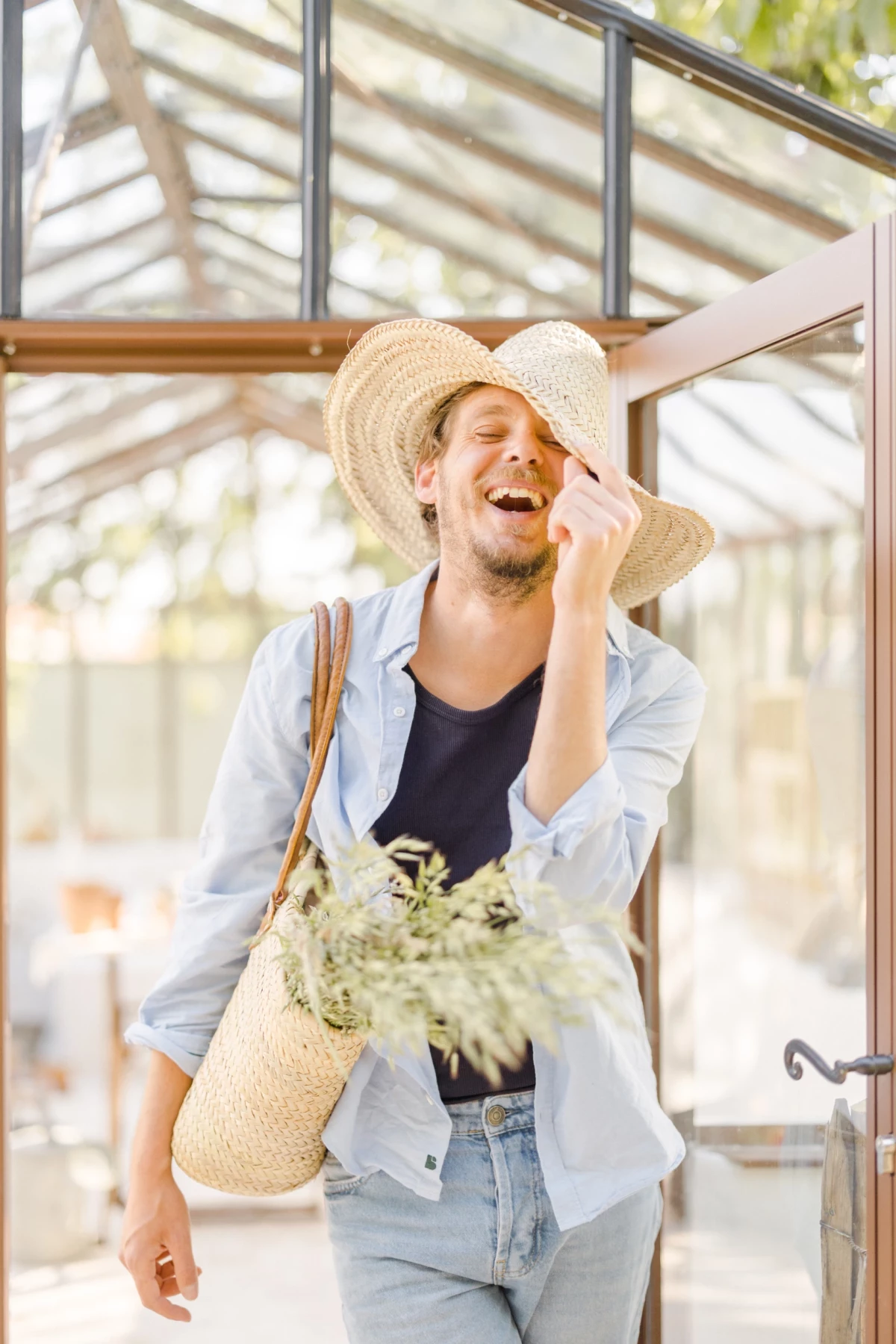 Portrait corporate fleuriste sous verrière : artisan souriant avec panier de fleurs et chapeau de paille, lumière naturelle douce