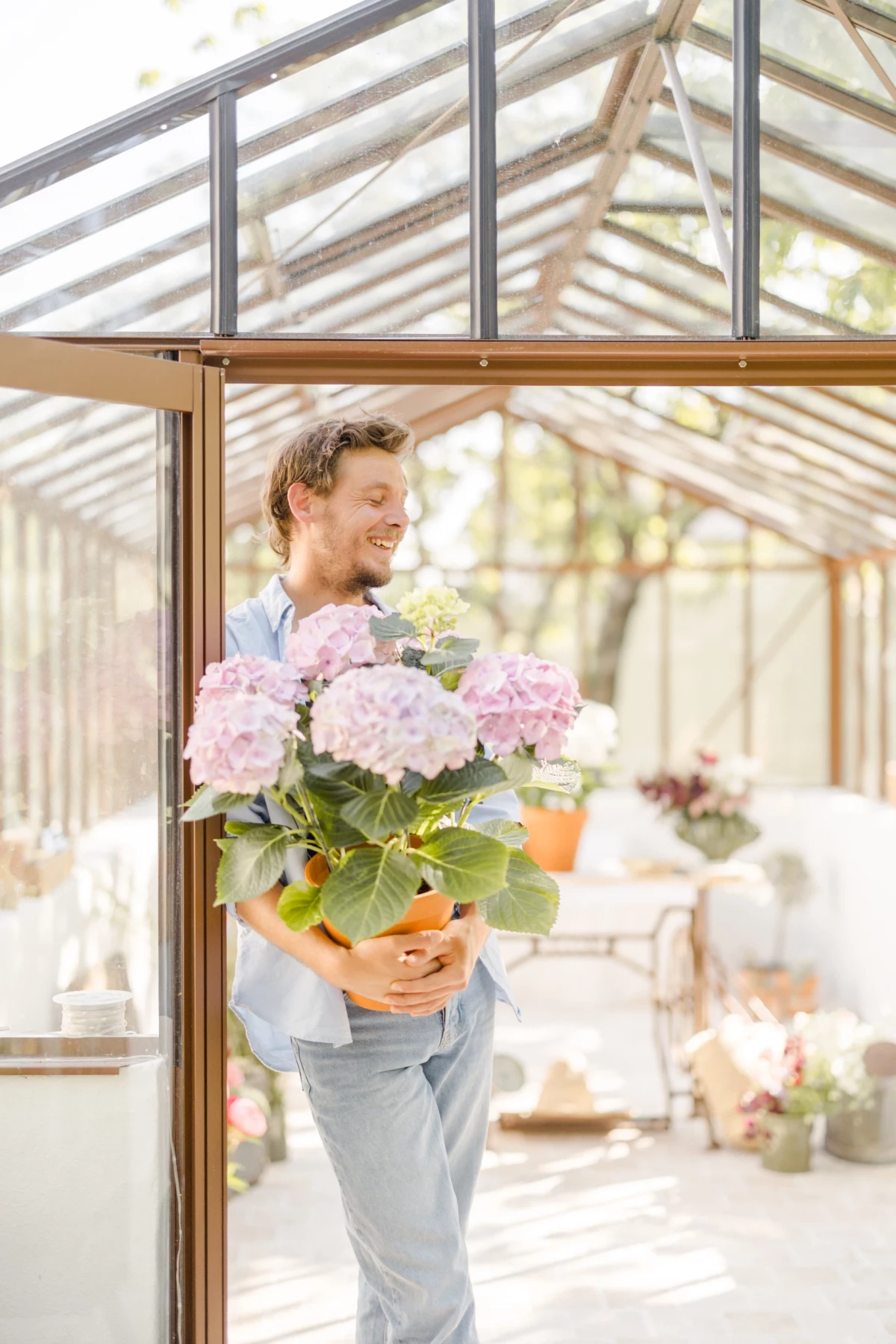 Portrait corporate fleuriste tenant un bouquet d'hortensias roses dans une verrière lumineuse, ambiance douce et professionnelle