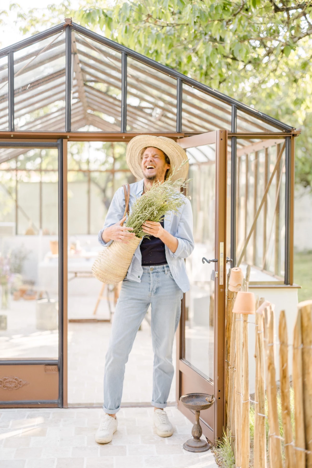 Portrait corporate fleuriste devant verrière : jeune femme souriante tenant bouquet champêtre et panier, ambiance lumineuse printanière