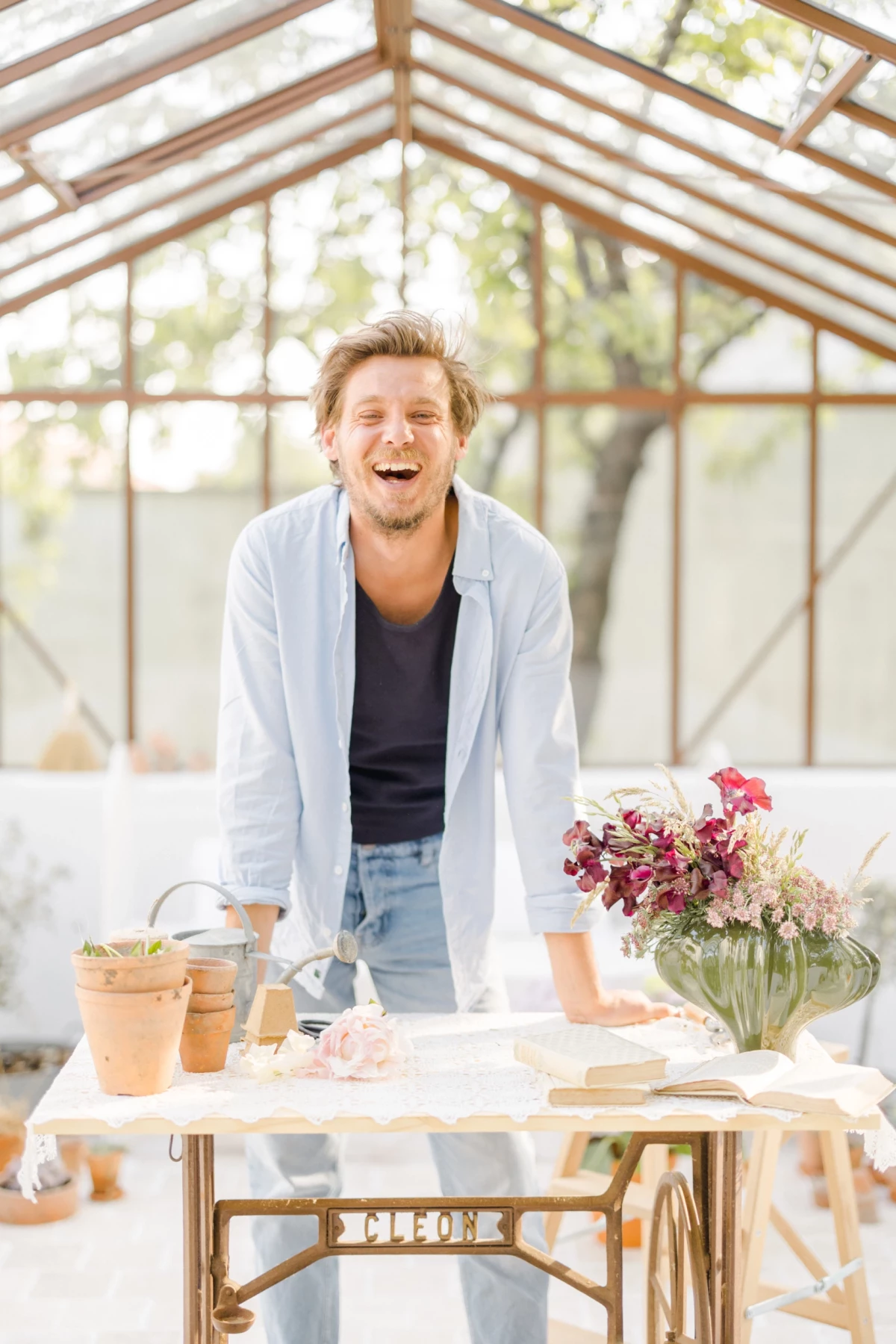 Portrait corporate fleuriste dans sa verrière : artisan souriant derrière sa table de travail avec bouquets et pots en terre