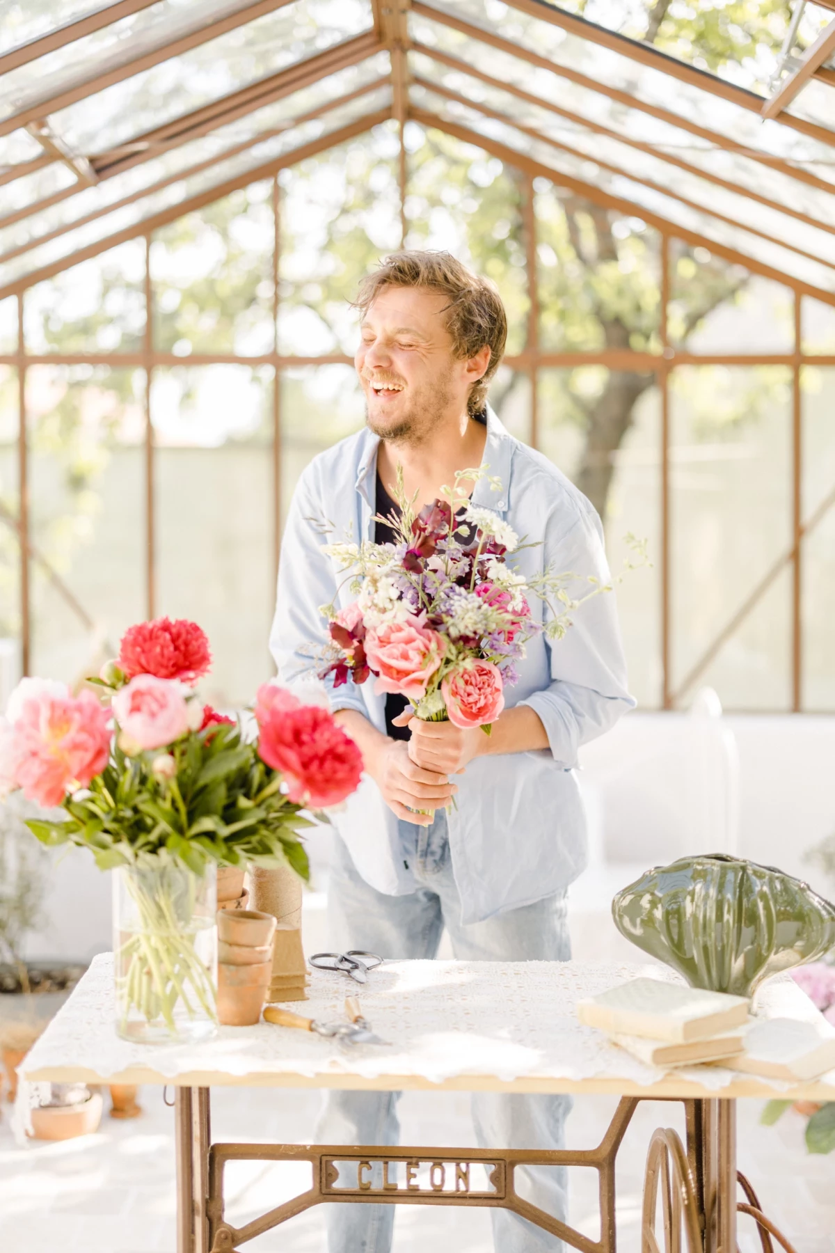 Portrait corporate fleuriste : artisan souriant arrange un bouquet de fleurs dans une verrière lumineuse en Vendée