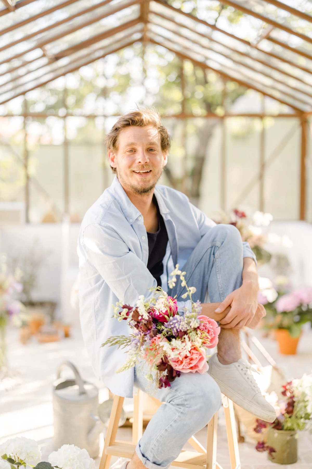 Portrait corporate d'un fleuriste souriant tenant un bouquet coloré dans une verrière lumineuse, ambiance printanière douce
