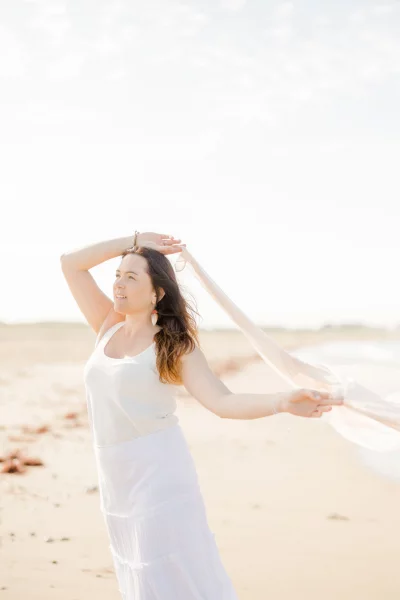 Femme en robe blanche sur une plage ensoleillée, levant les bras dans un geste de liberté et de joie