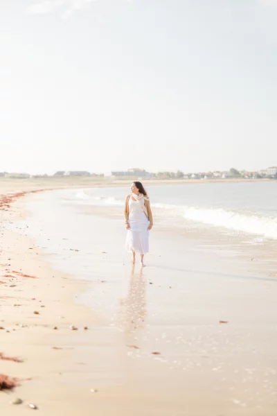 Femme en robe blanche marchant pieds nus sur une plage de sable au bord de l'eau
