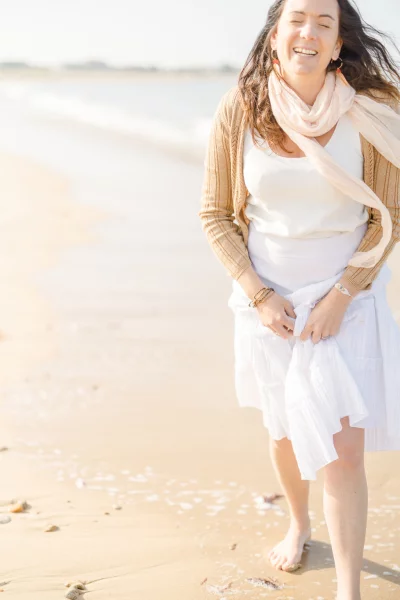 Femme souriante marchant pieds nus sur le sable d'une plage ensoleillée en robe blanche vaporeuse