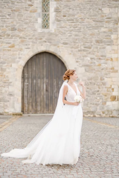 Mariée en robe blanche devant un château de pierre, voile au vent sur un sol pavé