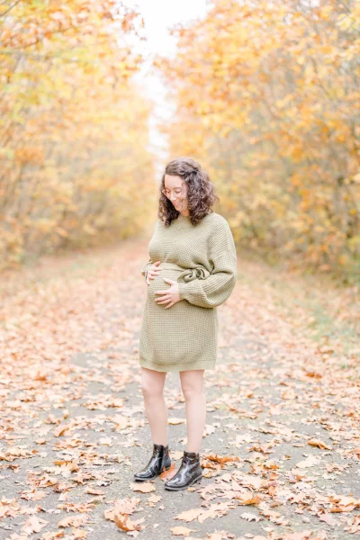Future maman en robe beige caressant son ventre rond dans une allée forestière parsemée de feuilles d'automne