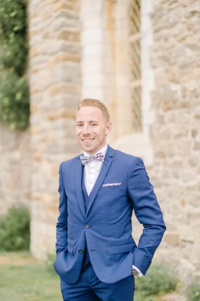 Marié souriant en costume bleu avec nœud papillon devant un château en pierre beige