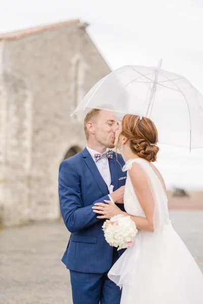Couple de mariés s'embrassant sous un parapluie transparent devant un château en pierre