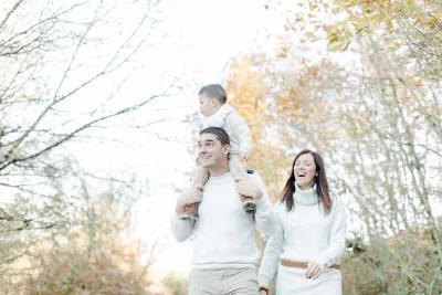 Famille complice en tenue blanche se promenant dans la nature automnale avec leur enfant sur les épaules
